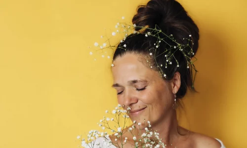 Man sieht eine Frau mit Schleierkraut im Haar und in den Händen. Sie hat die Augen geschlossen und steht vor einer gelb-orangenen Wand. Sie wirkt selig und versunken in etwas.